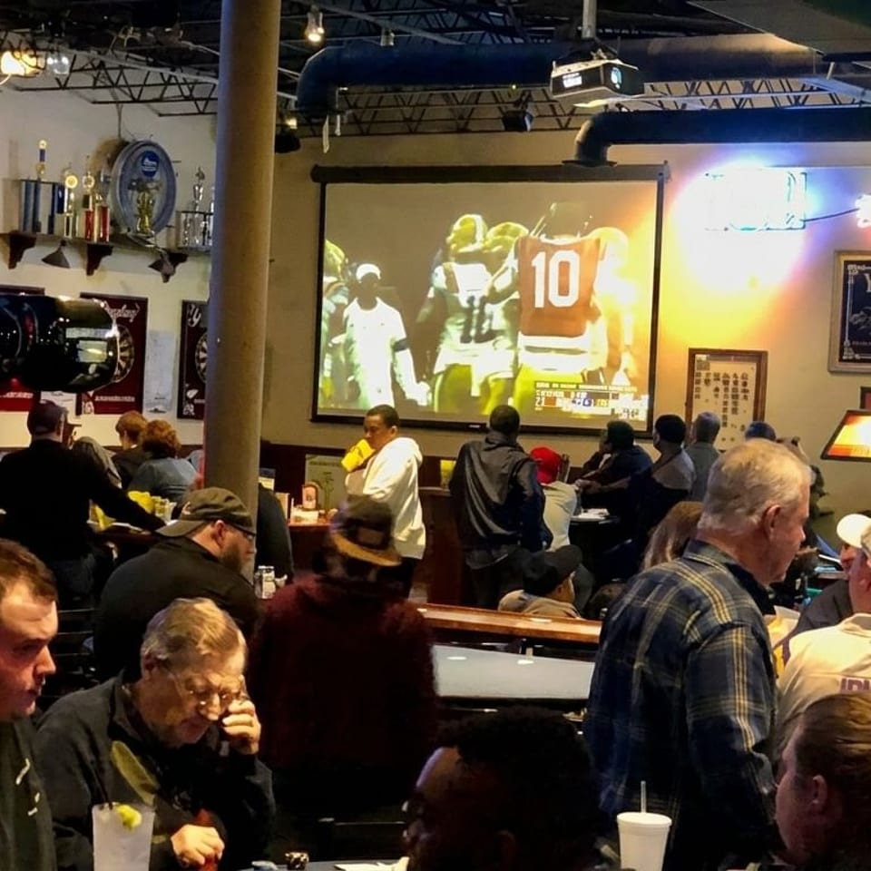 A lively bar scene with patrons watching a football game on a large projection screen.