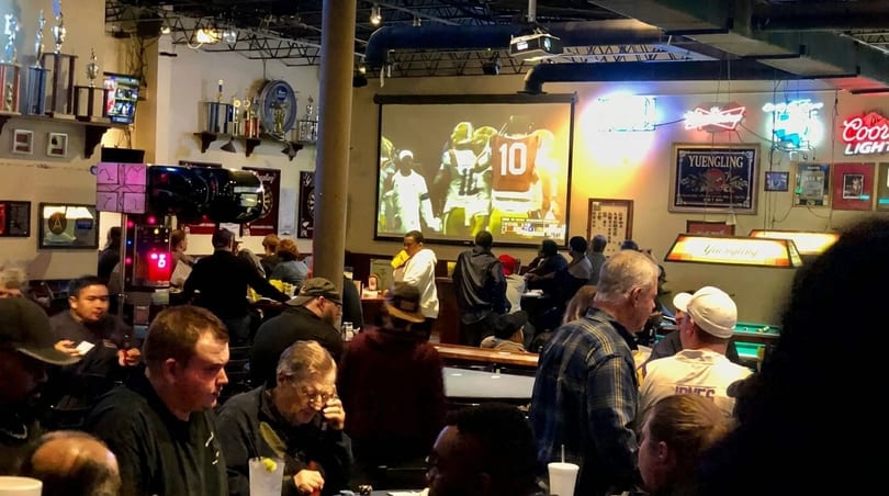 A lively bar scene with patrons watching a football game on a large projection screen.