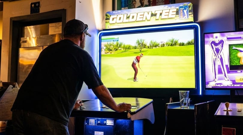 Man playing a Golden Tee golf arcade game in a dimly lit room.