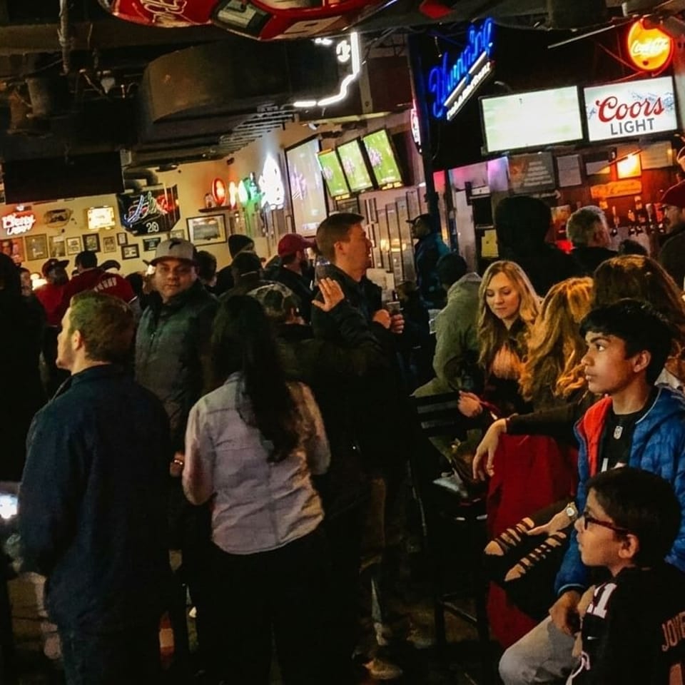 A crowded, dimly lit sports bar with multiple TVs showing a game and people socializing.