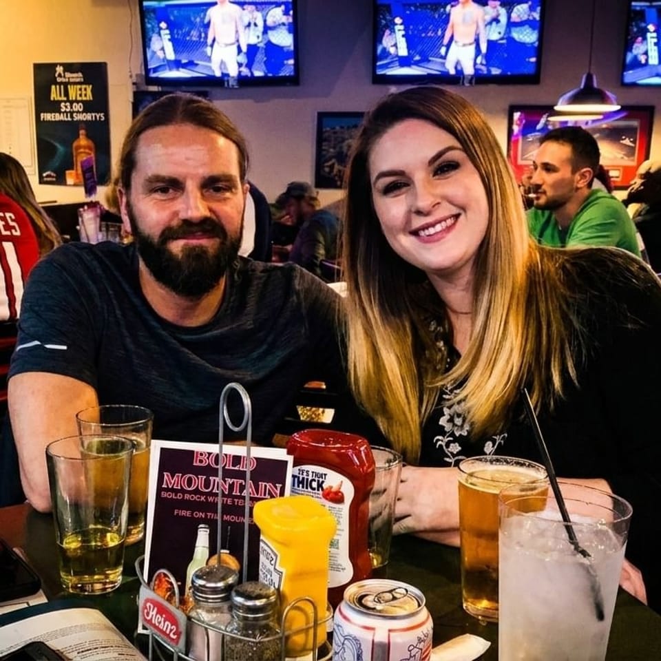 A smiling couple sits at a table in a lively bar with drinks and condiments, surrounded by other patrons.