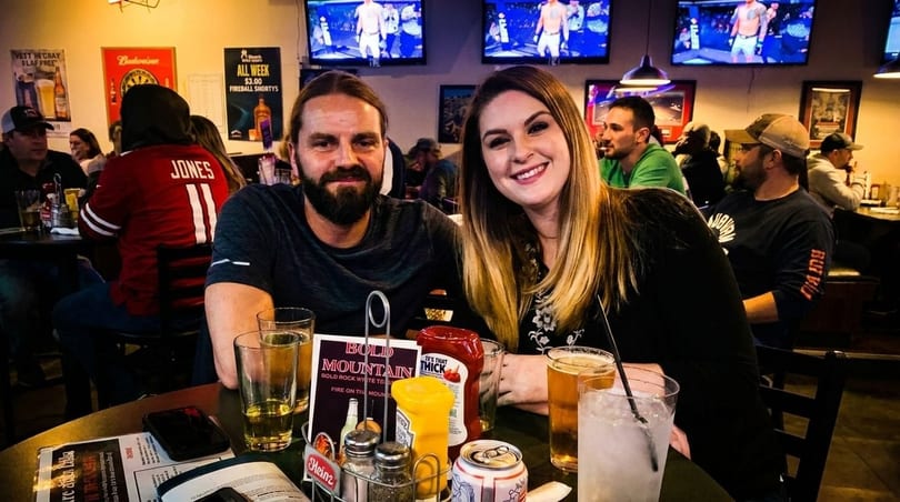 A smiling couple sits at a table in a lively bar with drinks and condiments, surrounded by other patrons.